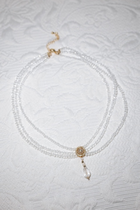 Wielorzędowa kolia z transparentnych kryształków ułożona na jasnej tkaninie w pełnej długości.